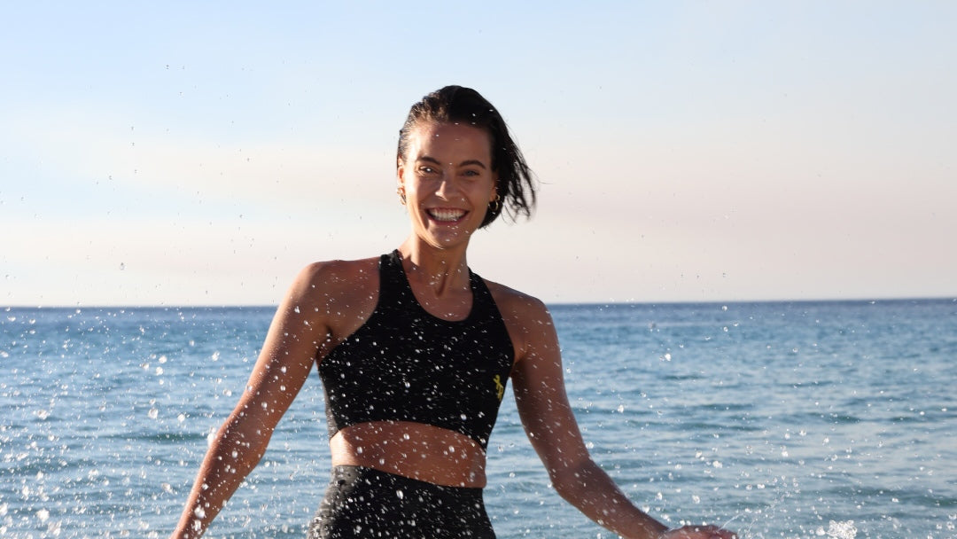 Woman in athletic black top standing by the ocean with water splashing around her.