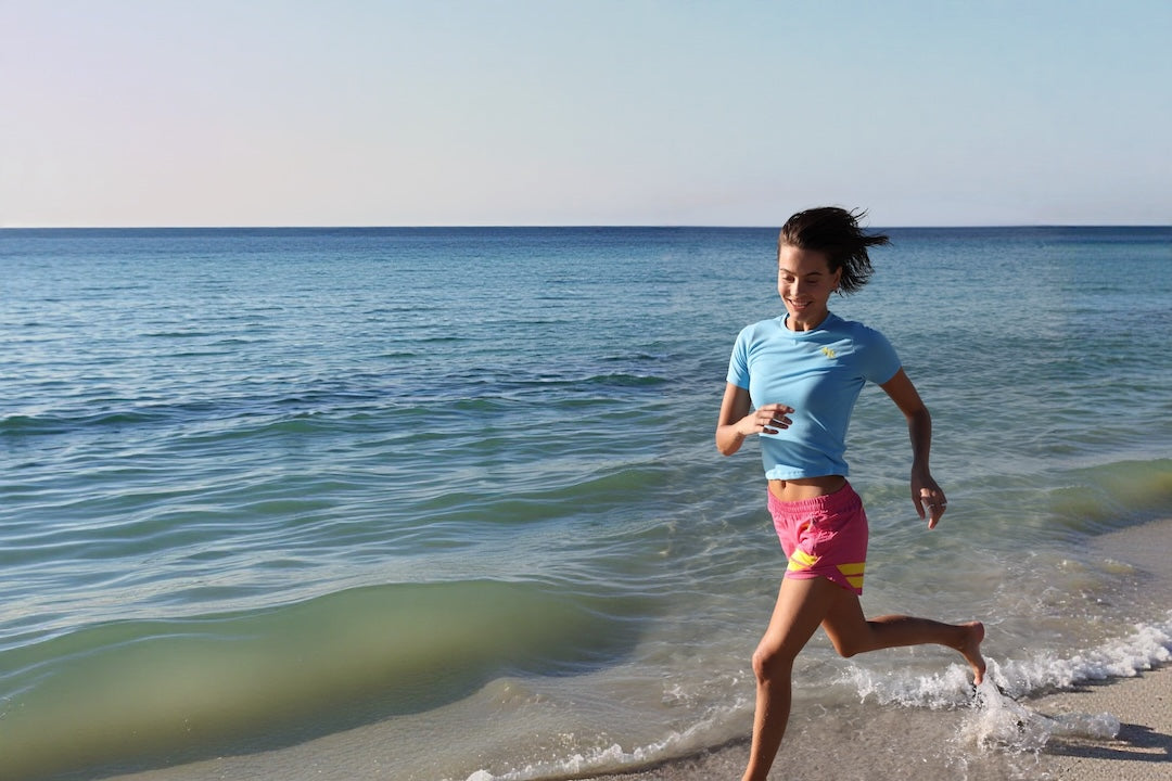 A woman running on a sandy beach shoreline wearing a light blue t-shirt and pink athletic shorts with yellow stripes by One More Reason premium sportswear for an active life