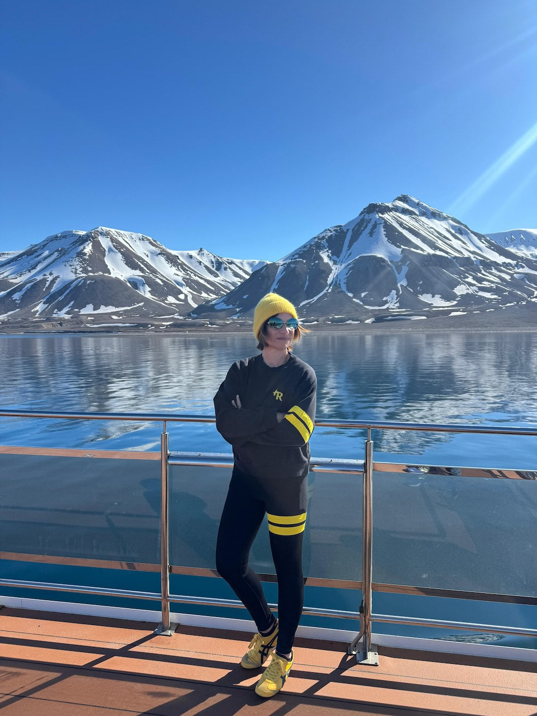 woman in a premium black sports outfit with yellow stripes from One More Reason standing on a boat deck with snow-covered mountains in the background