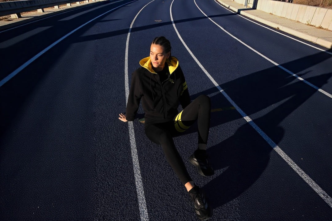 A woman sits on a blue running track wearing a black athletic hoodie with a yellow hood lining and matching black leggings with yellow stripes.