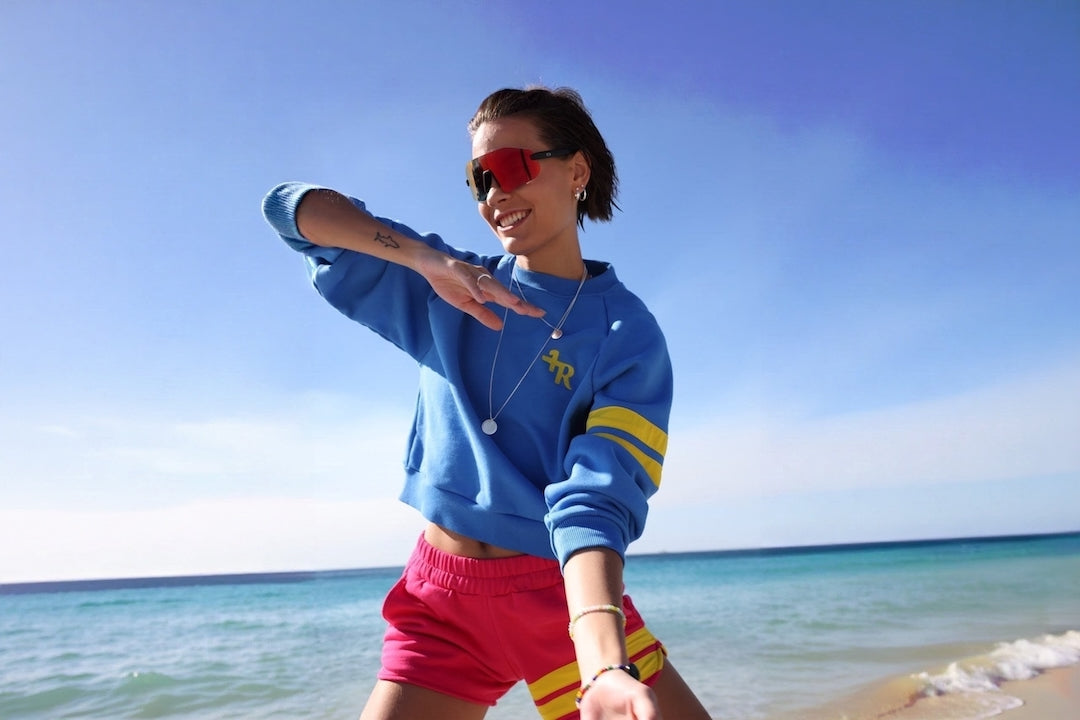 A woman posing on a beach wearing a blue cropped sweatshirt with yellow stripes on the sleeves, pink shorts, and red sunglasses.