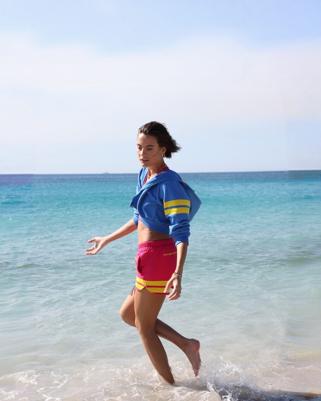 Woman in colorful sportswear outfit with yellow stripes by one more reason running on a beach with clear blue water and sky.