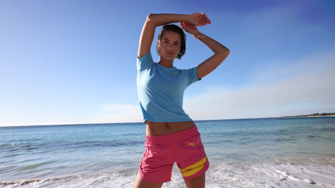 A woman posing on a beach wearing a light blue athletic t-shirt and pink athletic shorts with yellow stripes by one more reason, the premium sportswear brand crafted in Europe.