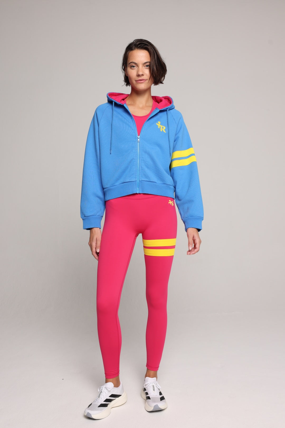 A model stands against a light gray studio background wearing a bright blue cropped zip-up hoodie with a pink hood lining and yellow stripes on the sleeves, paired with hot pink leggings featuring yellow stripes on the upper thigh and white sneakers. All by One More Reason, the premium women's sportswear brand.
