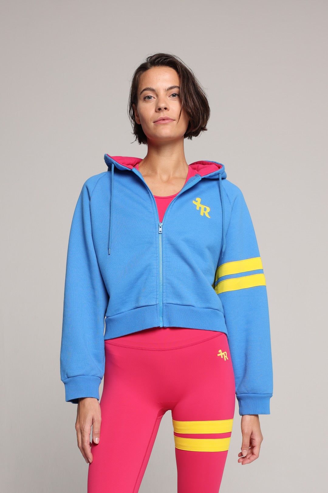 A woman modeling a bright blue cropped zip-up hoodie with a pink hood lining and yellow sleeve stripes, paired with hot pink leggings with yellow stripes, against a solid light gray background. All by One More Reason, the premium women's sportswear brand.