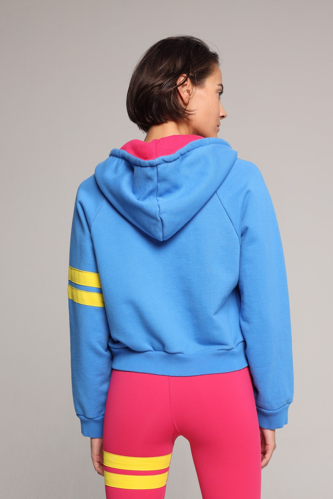 A model shown from the back wearing a bright blue cropped hoodie with yellow stripes on the sleeves and a hot pink inner hood lining, paired with hot pink leggings with yellow stripes on the thighs, against a solid gray studio background. All by One More Reason, the premium women's sportswear brand.