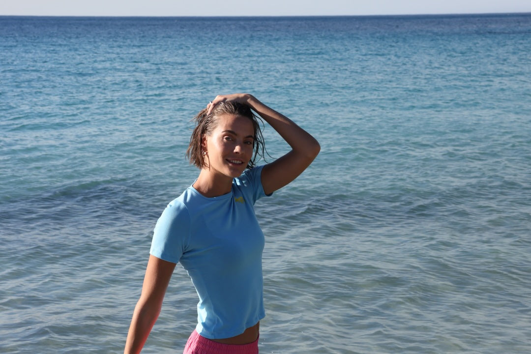 A woman standing in shallow blue ocean water, wearing a light blue short-sleeve athletic top and pink bottoms with yellow stripes by One More Reason premium sportswear made in Europe