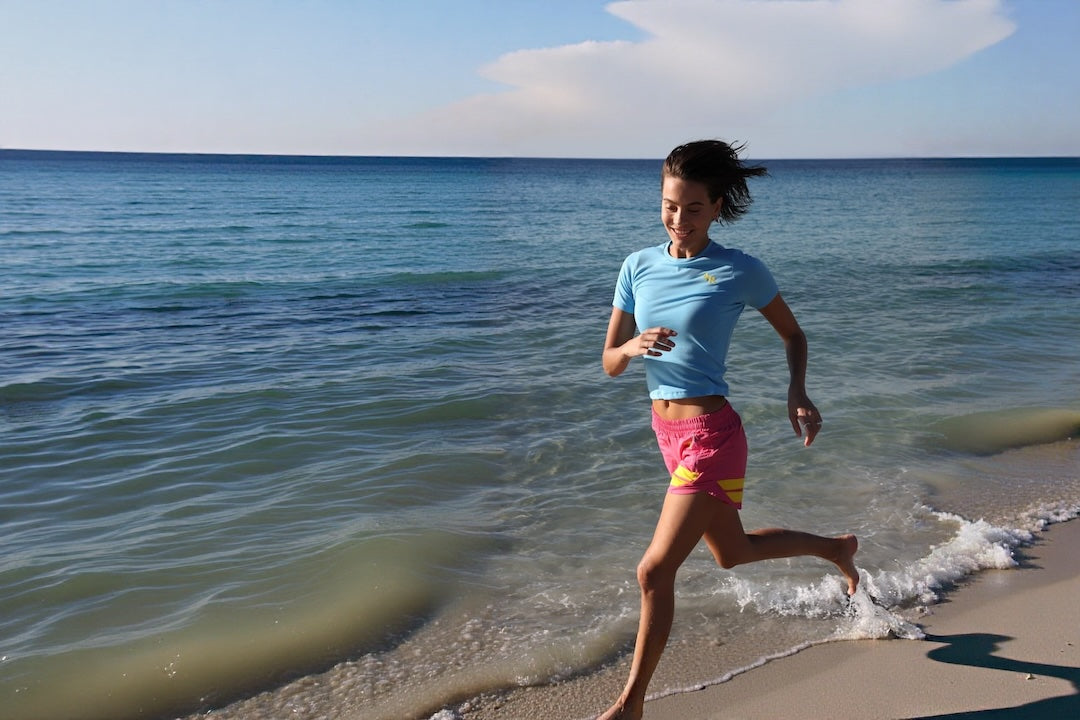 A woman running on a sandy beach shoreline wearing a light blue t-shirt and pink athletic shorts with yellow stripes by One More Reason premium sportswear for an active life