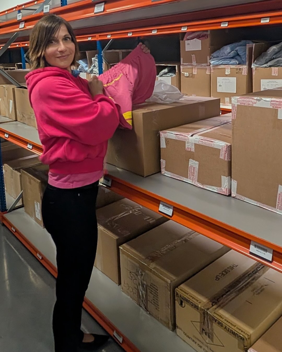 A woman in a bright pink hoodie and black pants stands in a warehouse aisle next to orange shelves filled with cardboard boxes, holding up a folded pink garment.