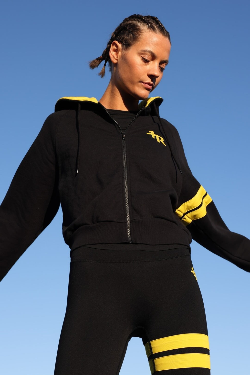 A woman wearing a black zip-up hoodie and matching black leggings with bright yellow accents, photographed outdoors against a clear blue sky. All from One More Reason, the European Premium Sportswear Brand.