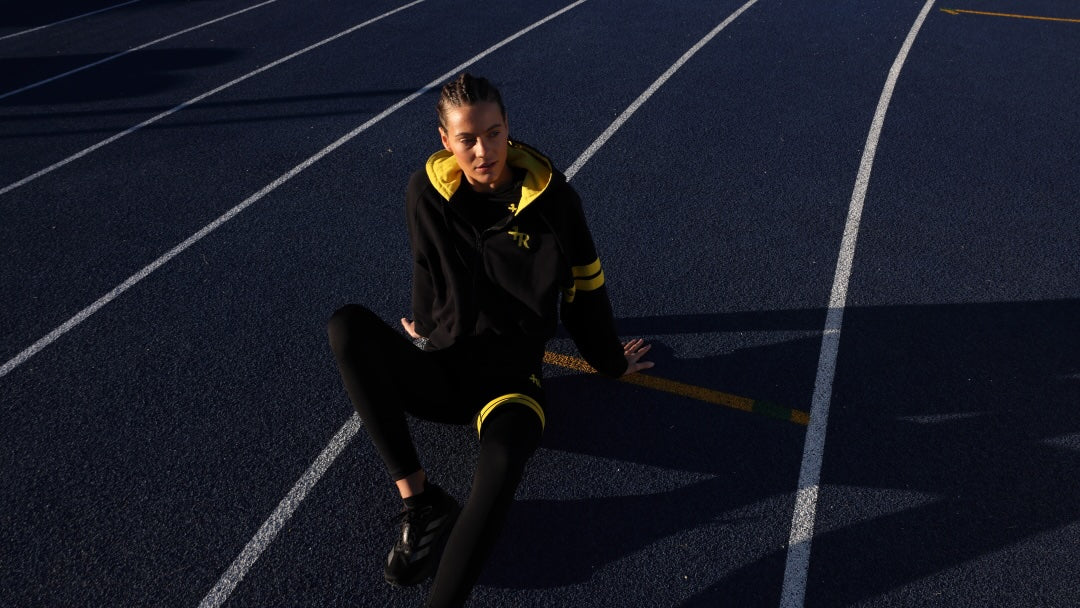 A female athlete sits on a dark blue athletic track field wearing a black athletic hoodie with bright yellow accents, black leggings, and black running shoes. All by One More Reason, the premium sportswear brand.
