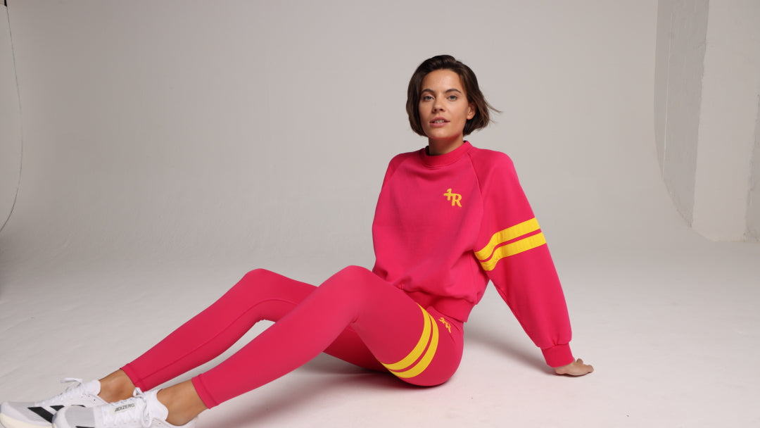 A woman wearing a bright pink two-piece tracksuit with yellow stripe accents sits on a light gray studio floor. The set includes a mock-neck sweatshirt with a yellow chest logo and matching leggings with yellow thigh stripes.