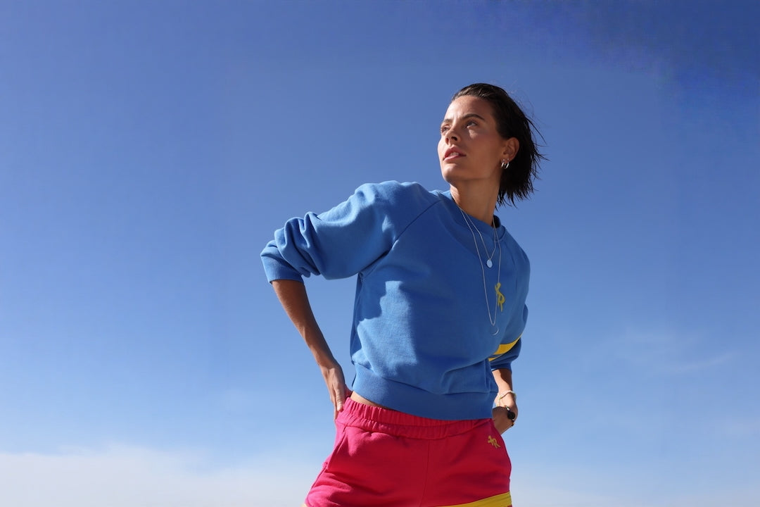 Woman wearing a blue sweatshirt with yellow stripes and pink shorts on a beach. All by one more reason, the premium sportswear brand, crafted in Europe.