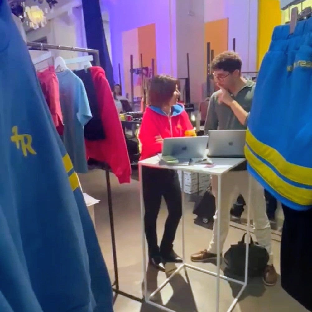 A lifestyle scene inside a clothing store or studio, showing a man and a woman standing behind a small white table with two laptops. Large blue athletic sweatshirts with yellow stripes are hanging on racks in the foreground, and other colorful apparel is visible in the background.