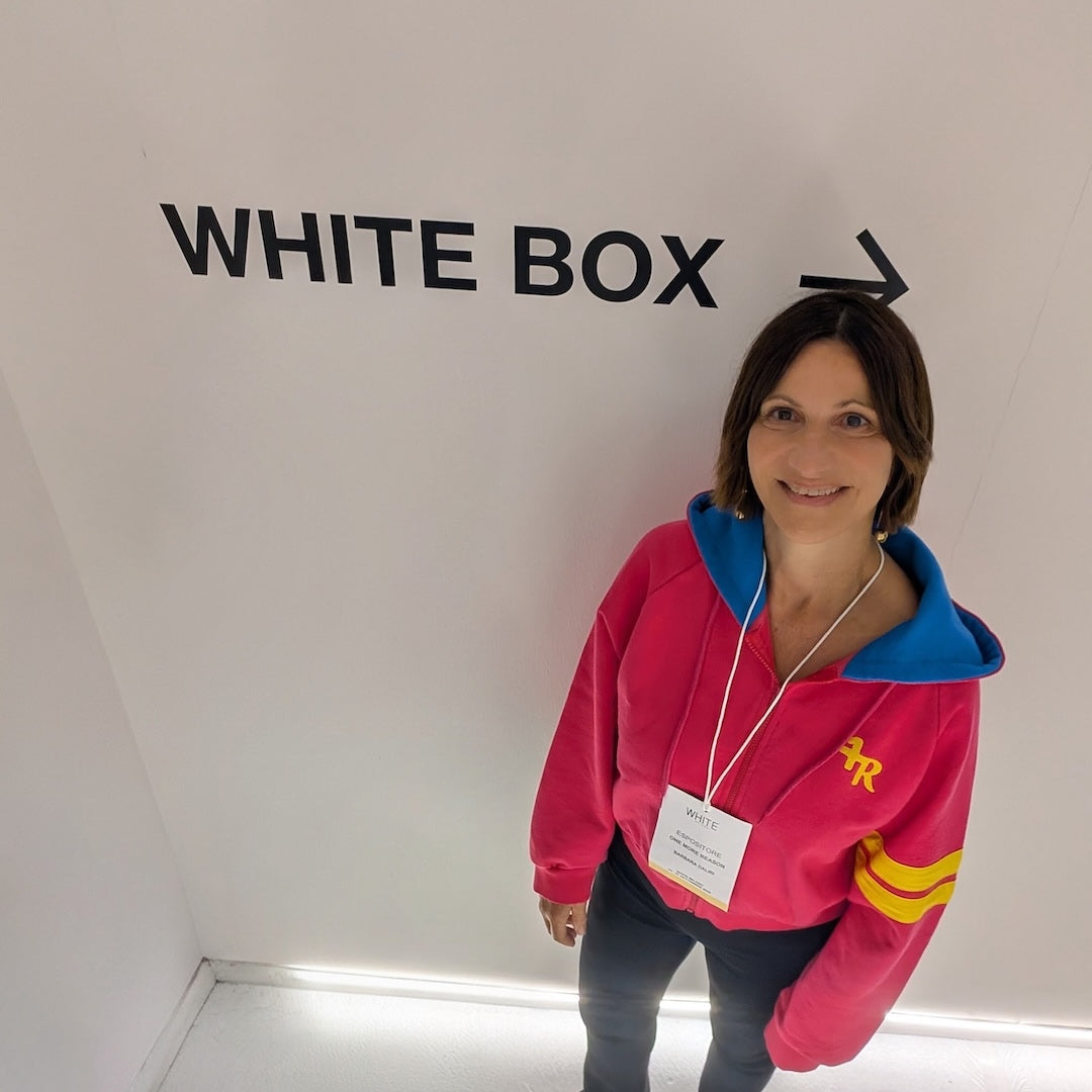 A woman wearing a bright pink hoodie with blue and yellow accents stands in front of a white wall with the words 'WHITE BOX' and an arrow pointing right.
