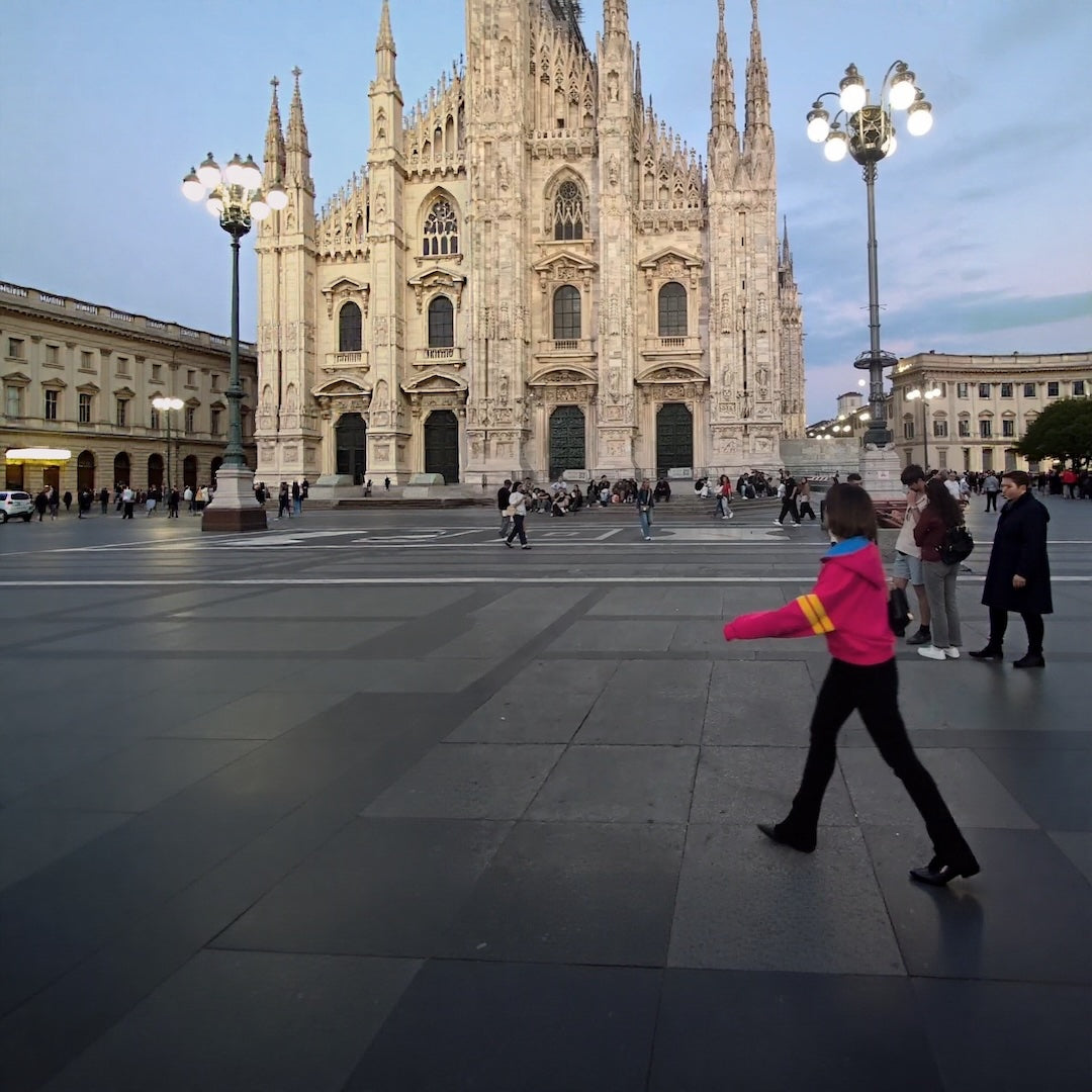 Woman in bright pink jacket with yellow stripes and black trousers walking in Milan in front of the Duomo.