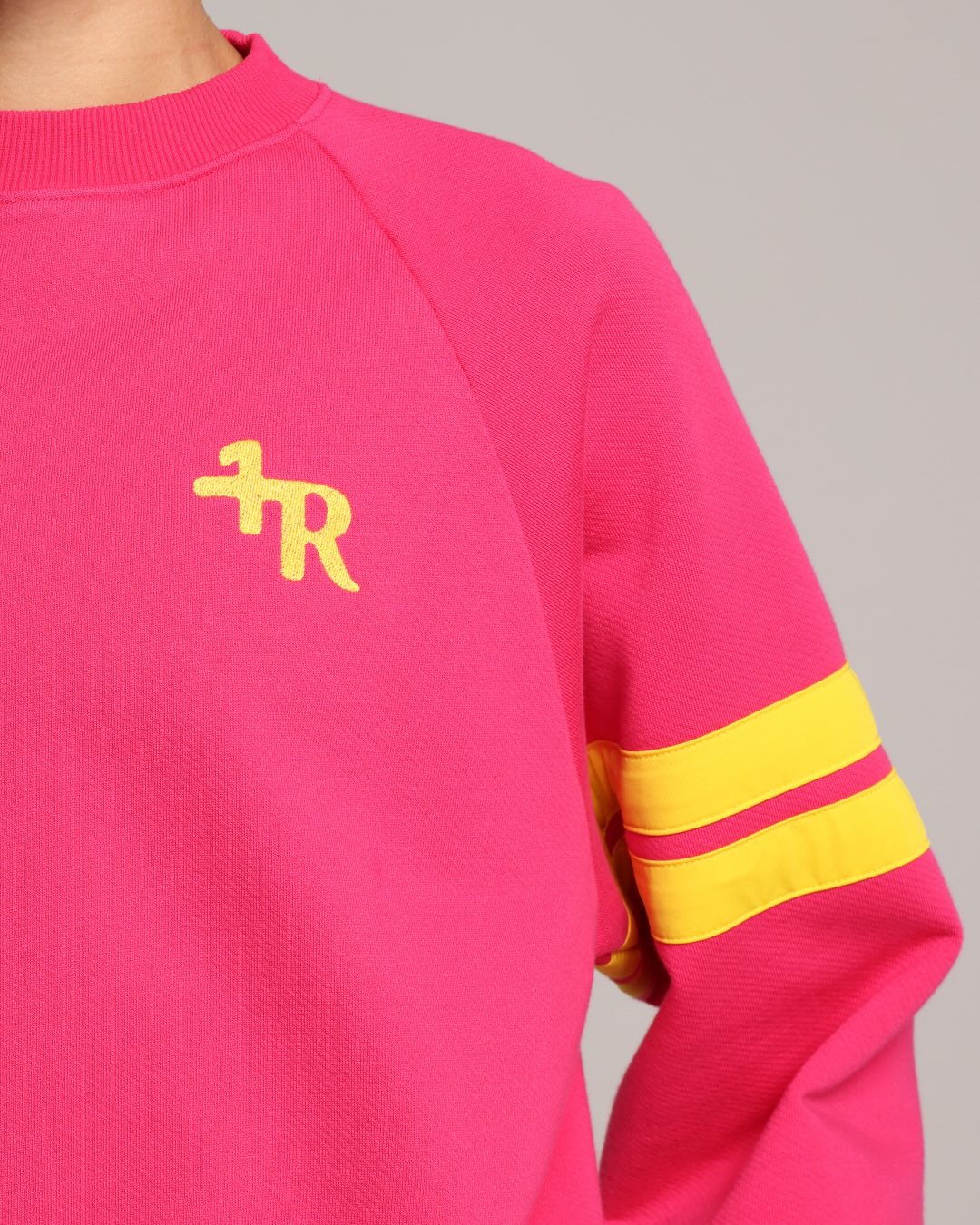 A close-up of a bright pink crewneck sweatshirt with a yellow  logo on the chest and two yellow stripes on the sleeve, shown on a model against a neutral gray background.