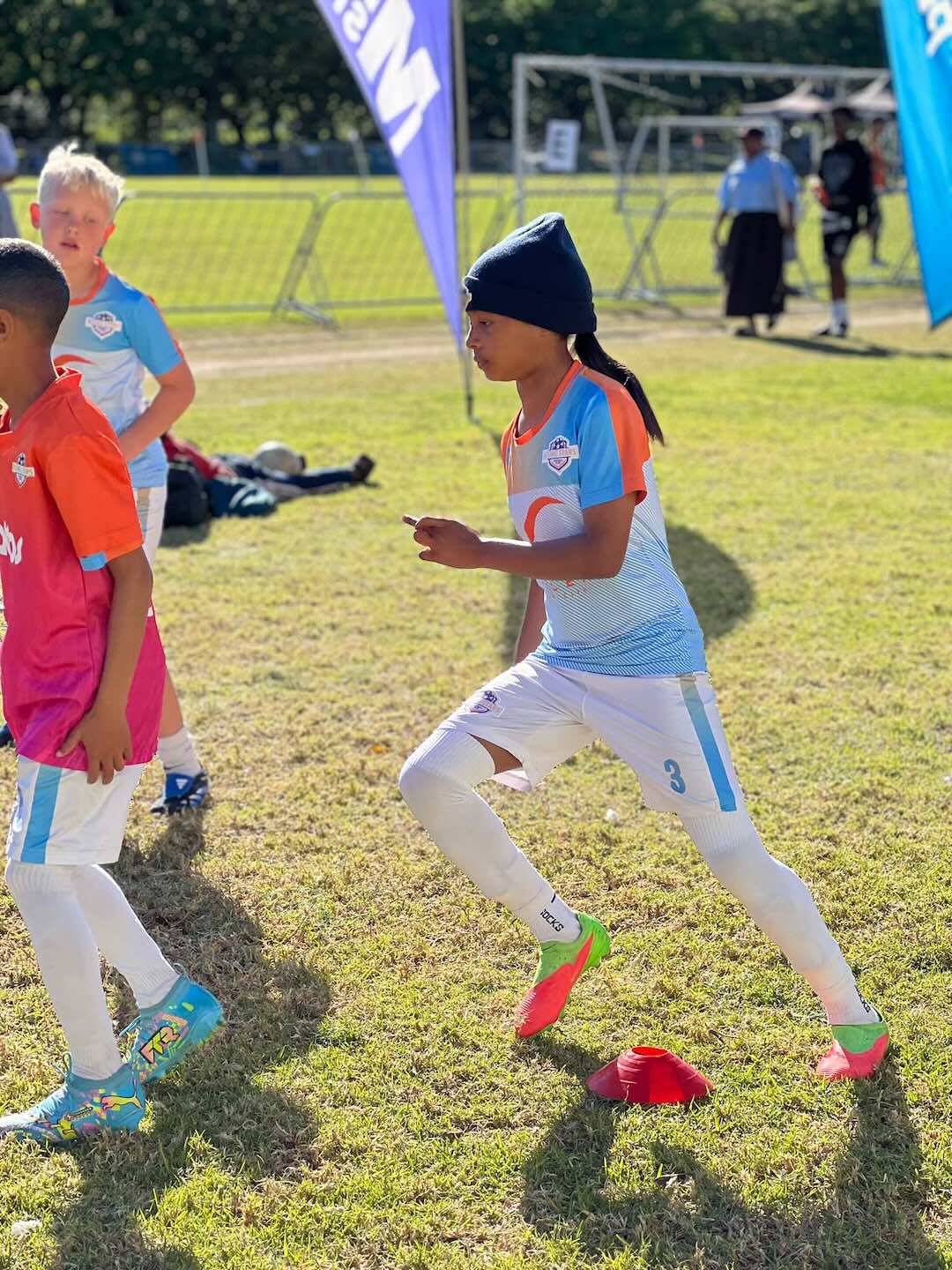 A youth soccer player in a light blue and orange jersey, white shorts, and a black beanie running on a grassy field, with other children and soccer equipment visible in the background.
