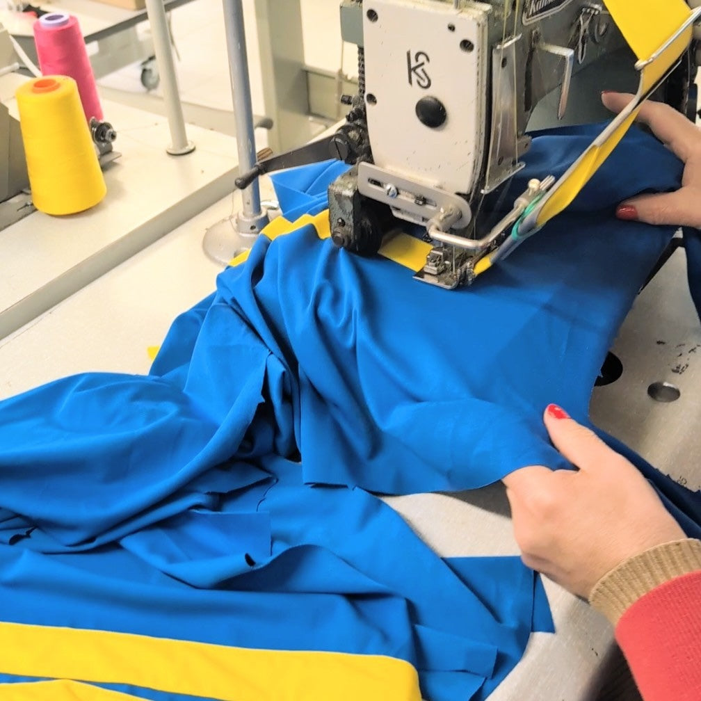 A close-up of a person guiding bright blue fabric with a yellow stripe under the presser bar of a white industrial sewing machine in a workshop setting.