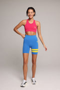 A woman in a bright pink sports bra and blue athletic biker shorts with yellow stripes, standing against a neutral gray studio background. Premium Sportswear by One More Reason.