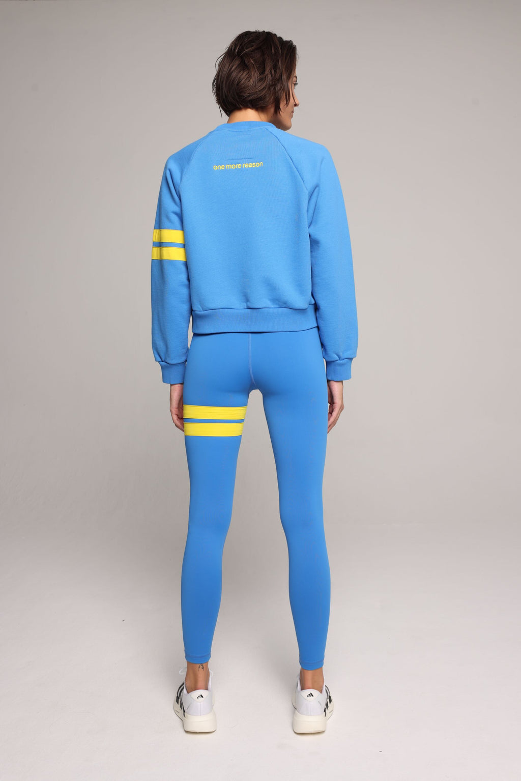 Back view of woman wearing blue high-waisted leggings and oversized  blue crew sweater with yellow stripes by One More Reason, studio shot.