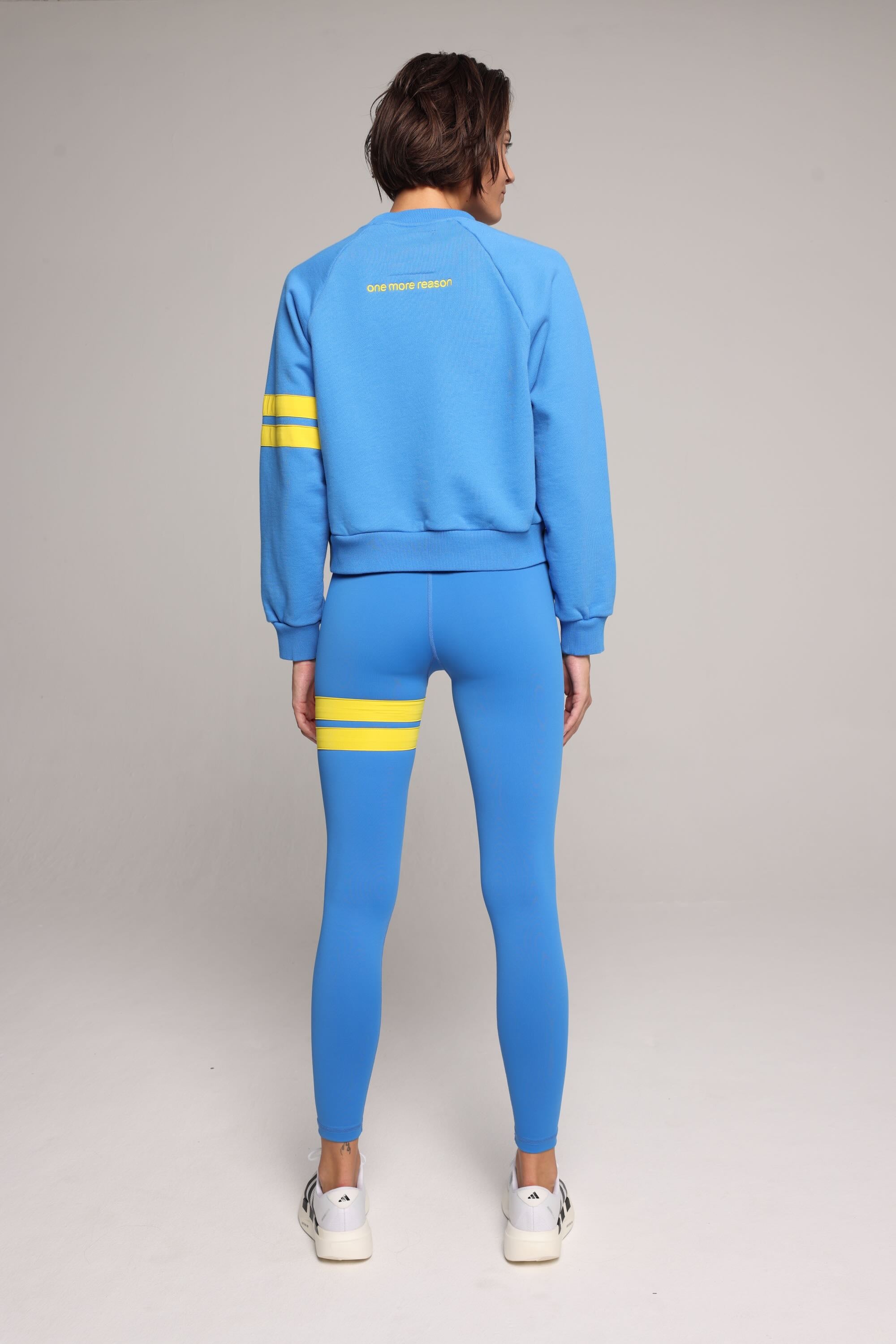 Back view of woman wearing blue high-waisted leggings and oversized  blue crew sweater with yellow stripes by One More Reason, studio shot.