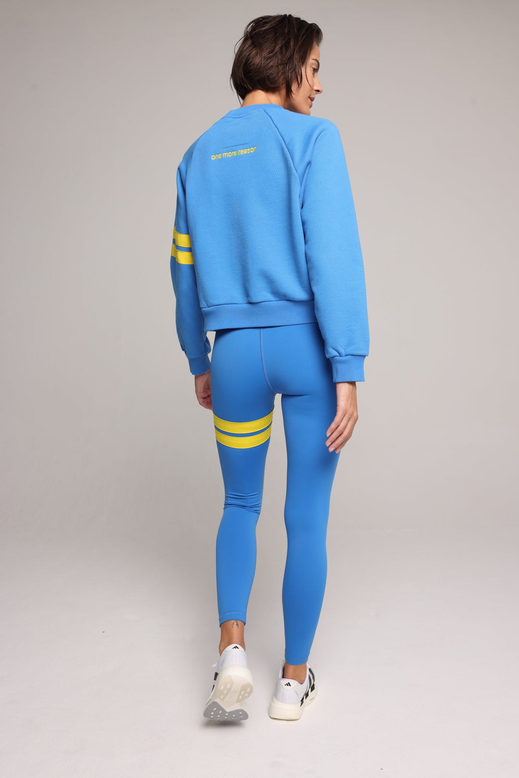 Back view of woman wearing blue high-waisted leggings and oversized  blue crew sweater with yellow stripes by One More Reason, studio shot.