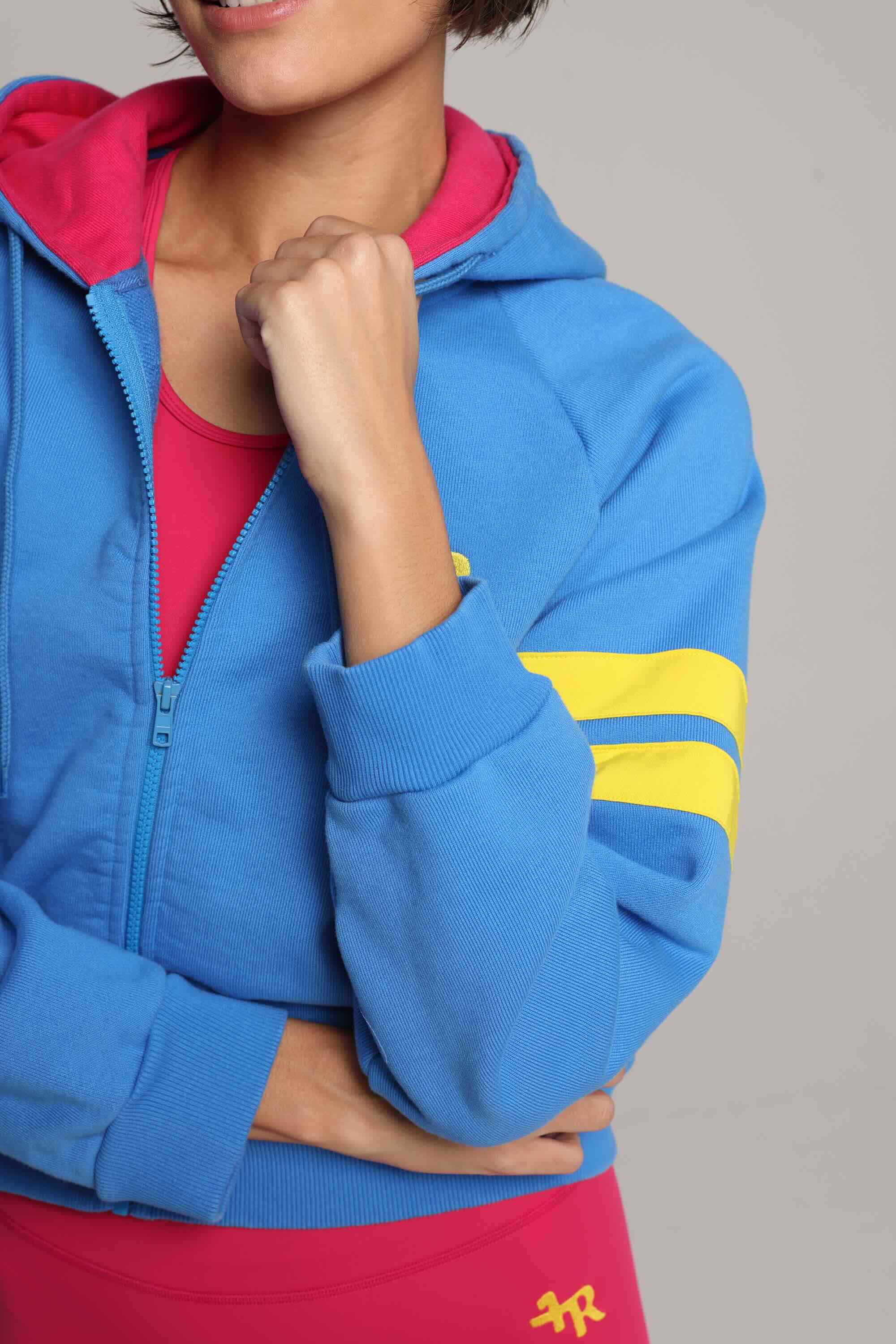 Woman wearing blue and pink luxury athletic set, blue bicolor zip hoodie by  One More Reason, with yellow stripes, studio look.