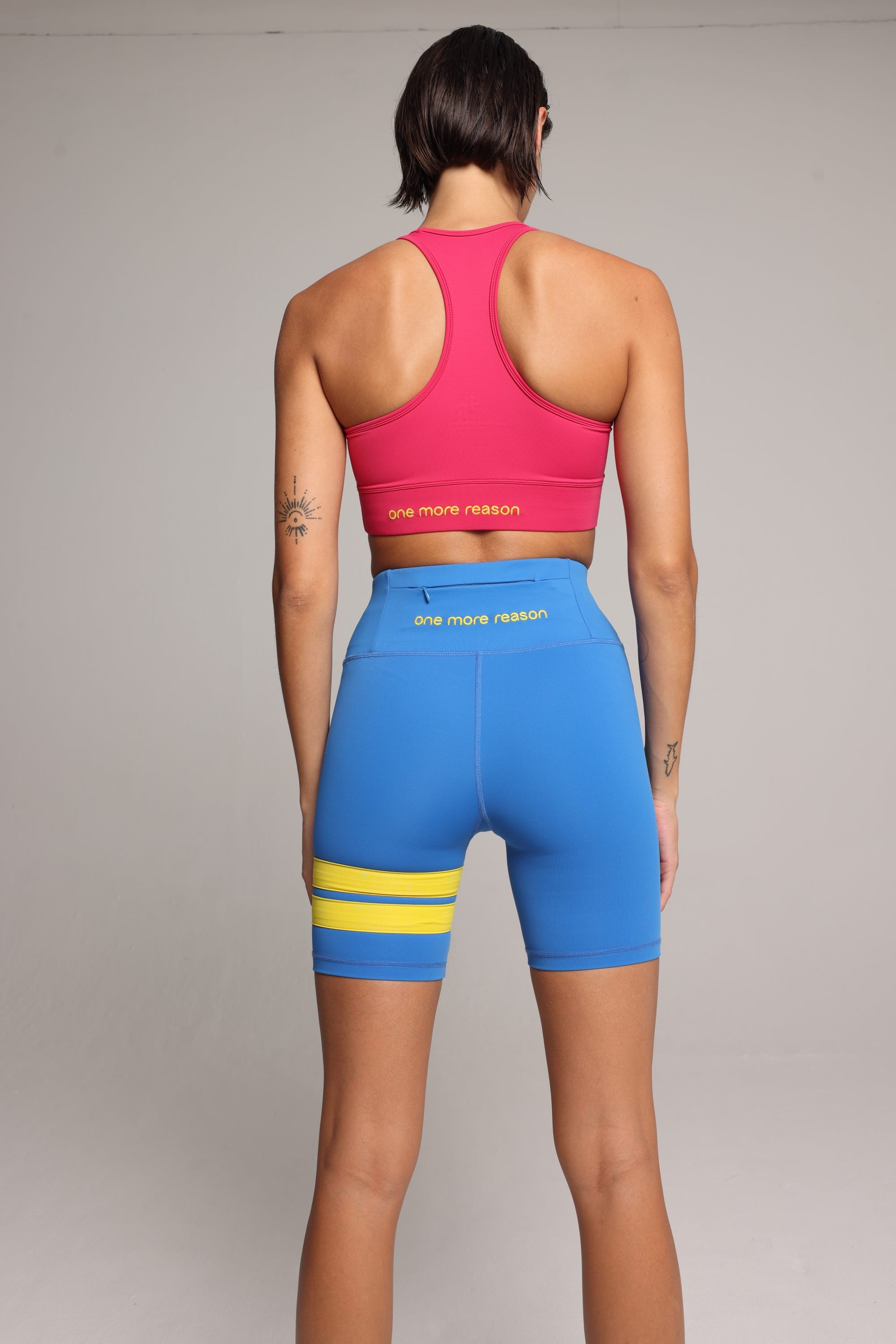 Woman wearing a soft pink sports bra and blue short leggings tights with yellow stripes by One More Reason fashion forward luxury sports wear seen from the back