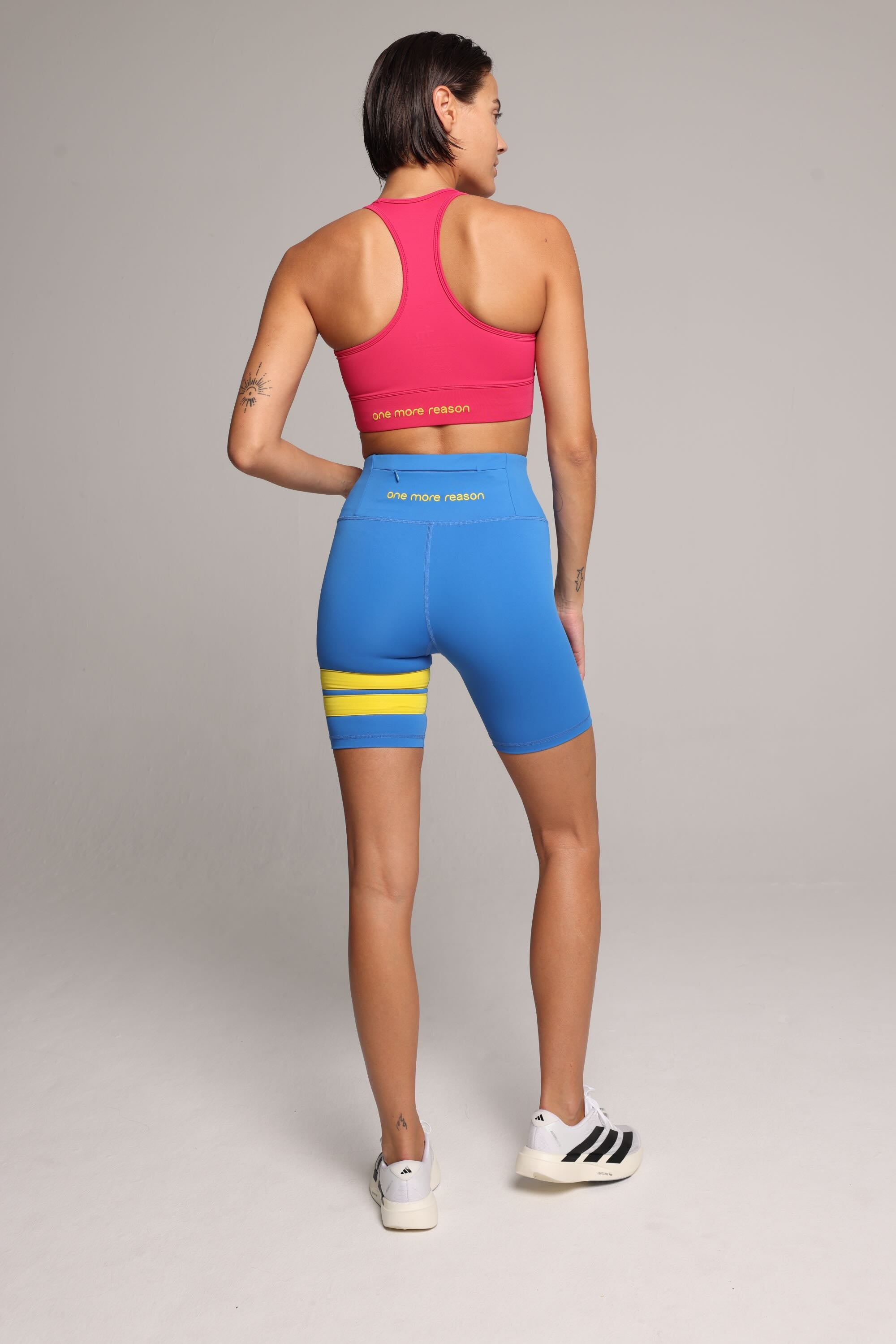 Woman wearing a soft pink sports bra and blue short leggings tights with yellow stripes by One More Reason fashion forward luxury sports wear seen from the back