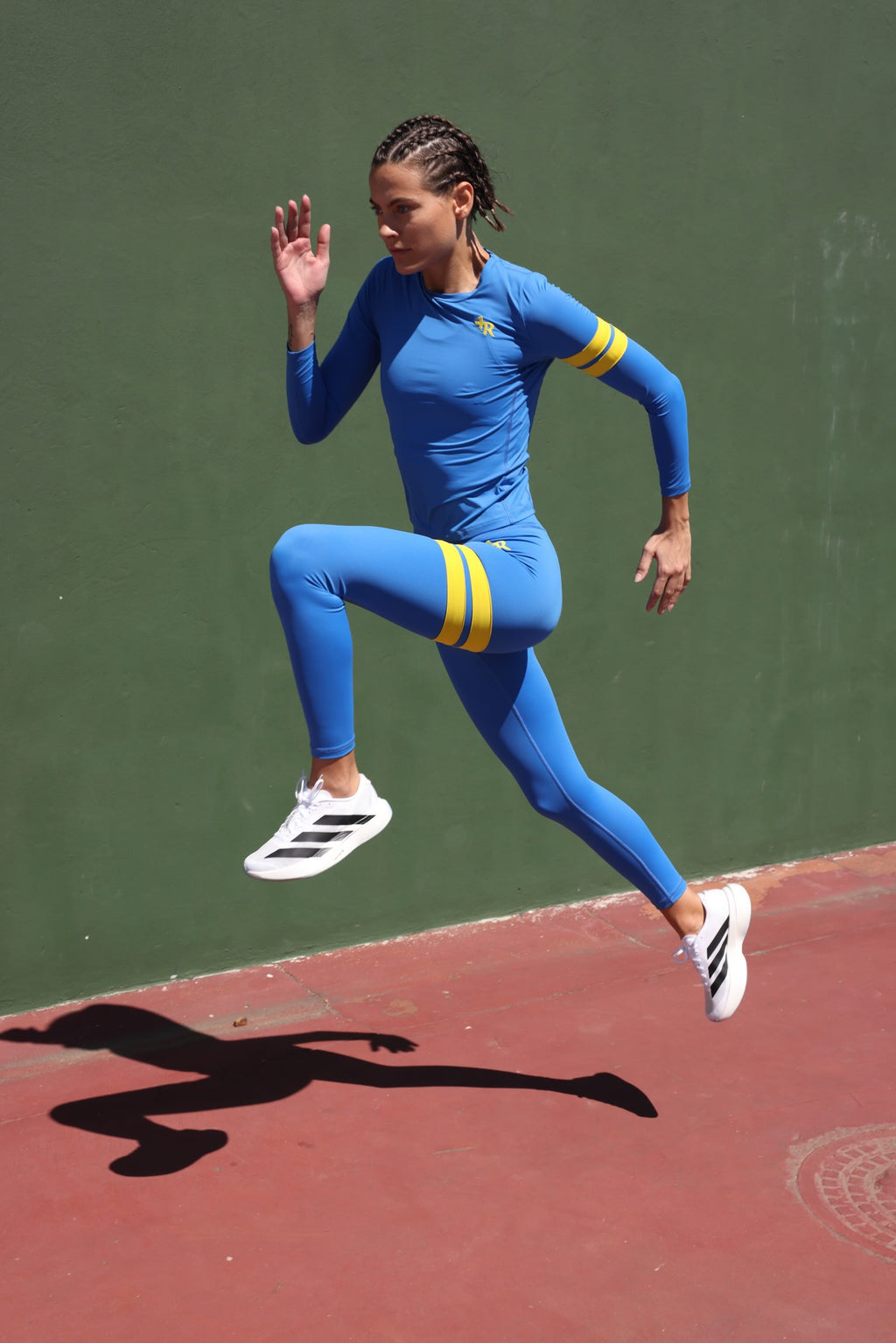 Women wearing premium blue high-waisted leggings and long-sleeve running top with yellow stripes by One More Reason fashion forward luxury sports wear, mid-jump on outdoor track