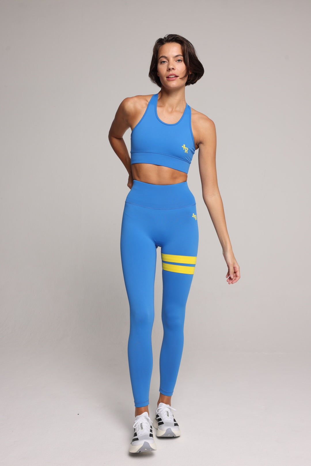 A woman modeling a bright blue athletic set consisting of a sports bra and high-waisted leggings with yellow stripe accents, standing against a neutral gray studio background. All by One More Reason, the premium women's sportswear brand.