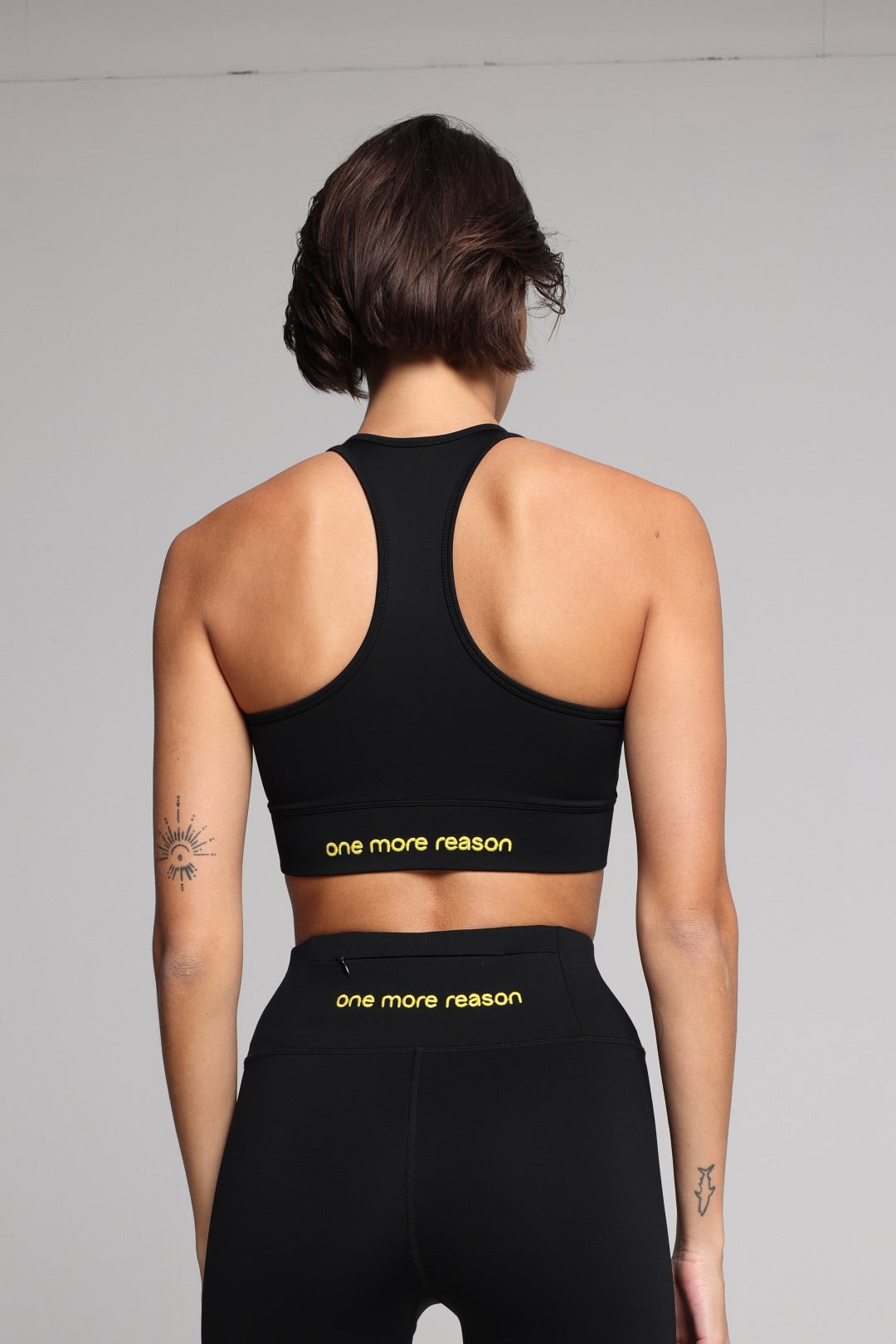 Rear view of a woman wearing a black sports bra and matching high-waisted leggings with yellow 'one more reason' branding on the underbust band, set against a plain light gray studio background. All by One More Reason, the premium women's sportswear brand.