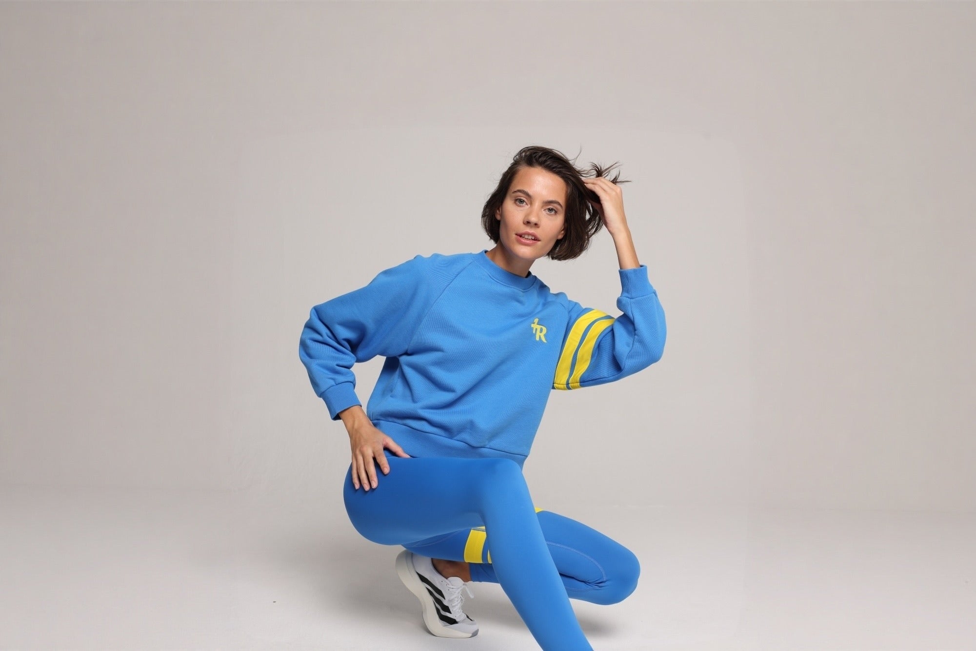 A woman posing in a bright blue athletic tracksuit with yellow stripes on the sleeves and thighs, paired with white sneakers, against a neutral light gray studio background. All by One More Reason, the premium sportswear brand, crafted in Europe.