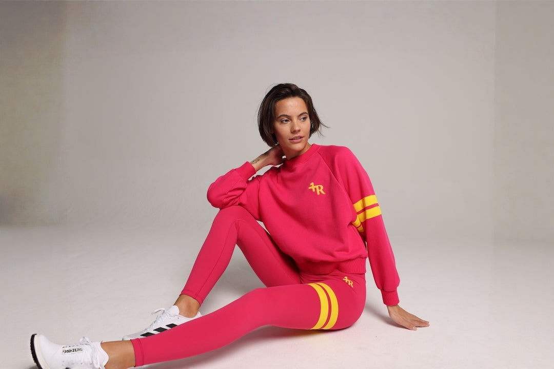 A woman sits on a light gray studio floor wearing a bright pink matching tracksuit with yellow stripe accents and white sneakers. All by One More Reason, the premium sportswear brand, crafted in Europe.