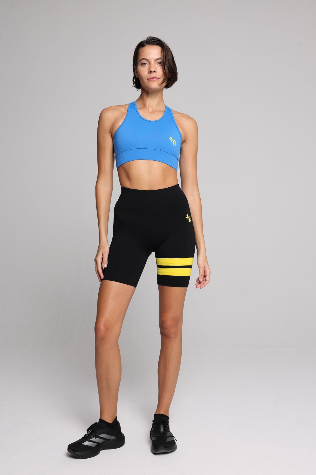 A woman in a bright blue athletic sports bra and black biker shorts with yellow stripes, standing against a light gray studio background. Premium Sportswear by One More Reason.