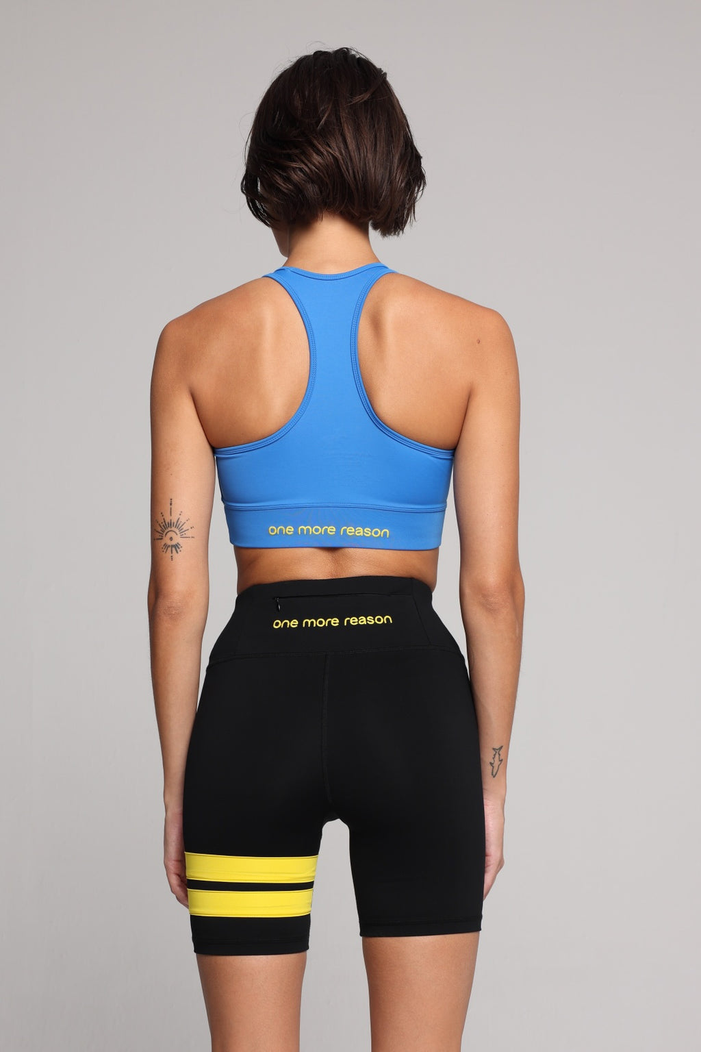 A woman shown from the back wearing a bright blue racerback sports bra and black biker shorts with yellow stripes and yellow text reading 'one more reason' on a light gray studio background. Premium Sportswear by One More Reason.
