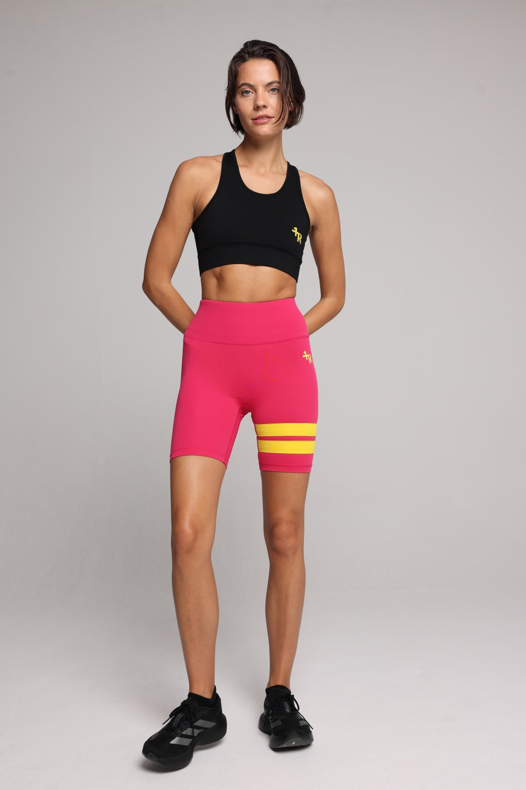 A woman in a pink athletic biker shorts with two yellow stripes on the lower right leg, paired with a black sports bra and black sneakers, against a light gray studio background. Premium Sportswear by One More Reason.
