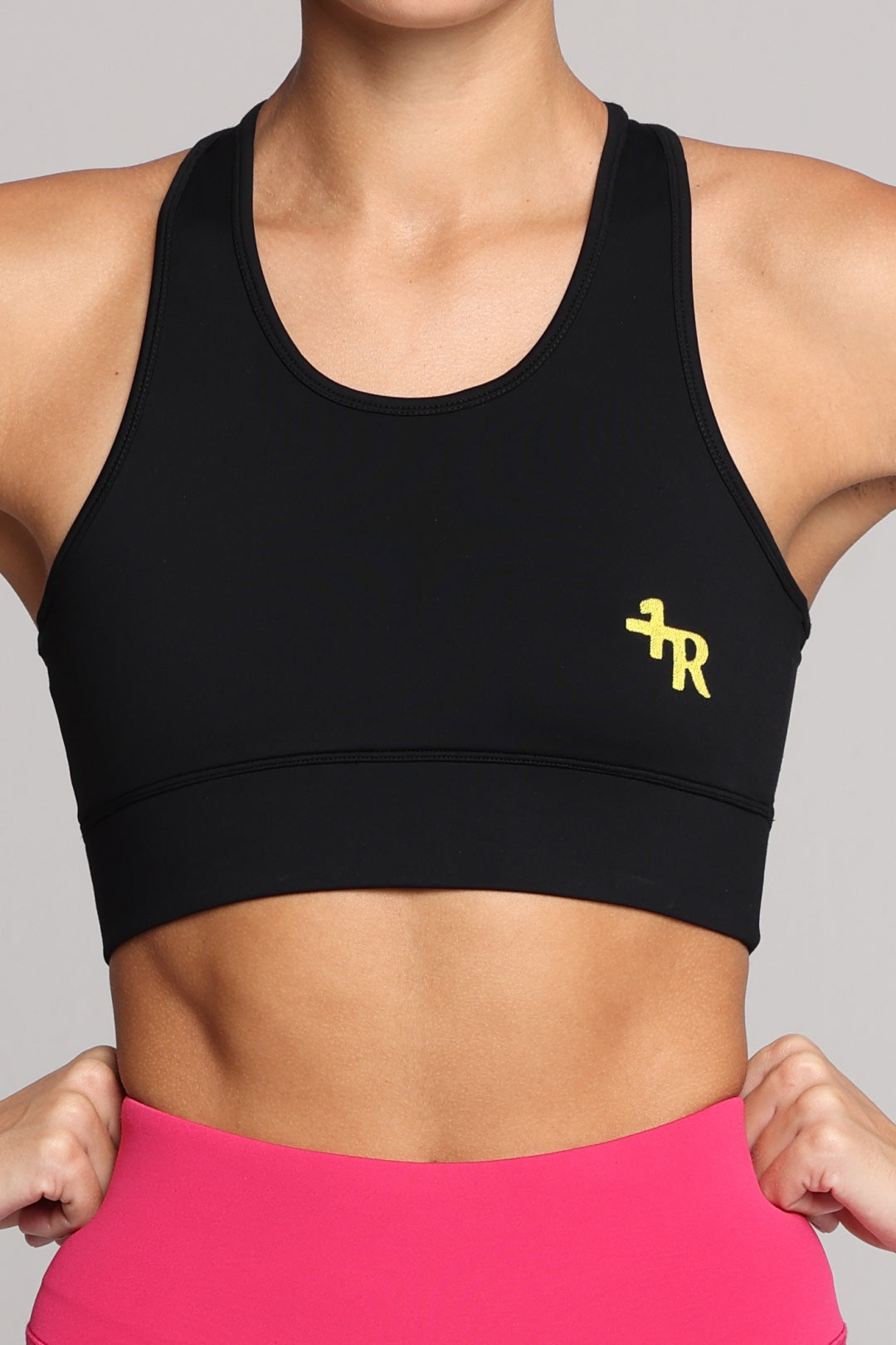 A close-up of a woman wearing a black athletic sports bra with a small yellow logo on the left chest, paired with bright pink bottoms, against a solid gray background. Premium Sportswear by One More Reason.