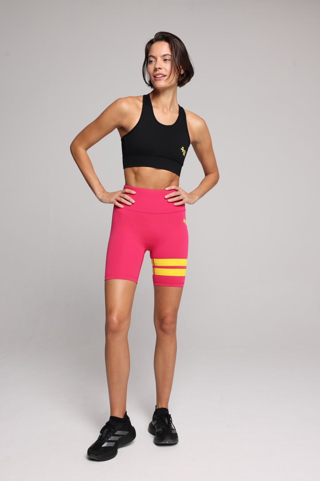 A woman in bright pink athletic biker shorts with two yellow stripes near the hem, paired with a black sports bra and black sneakers, against a light gray studio background. Premium Sportswear by One More Reason.