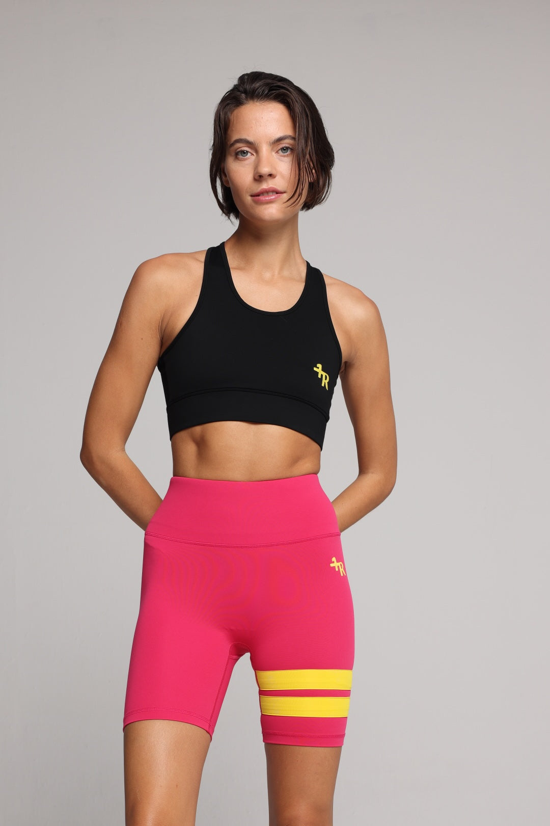 A woman in a black sports bra and bright pink biker shorts with yellow stripes, photographed against a neutral gray studio background. Premium Sportswear by One More Reason.