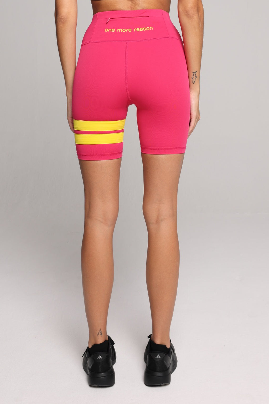 Rear view of a woman wearing bright pink biker shorts with two horizontal yellow stripes on the left leg. Premium Sportswear by One More Reason.