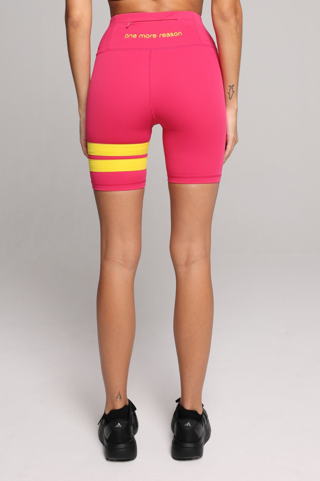 Rear view of a woman wearing bright pink biker shorts with two horizontal yellow stripes on the left leg. Premium Sportswear by One More Reason.