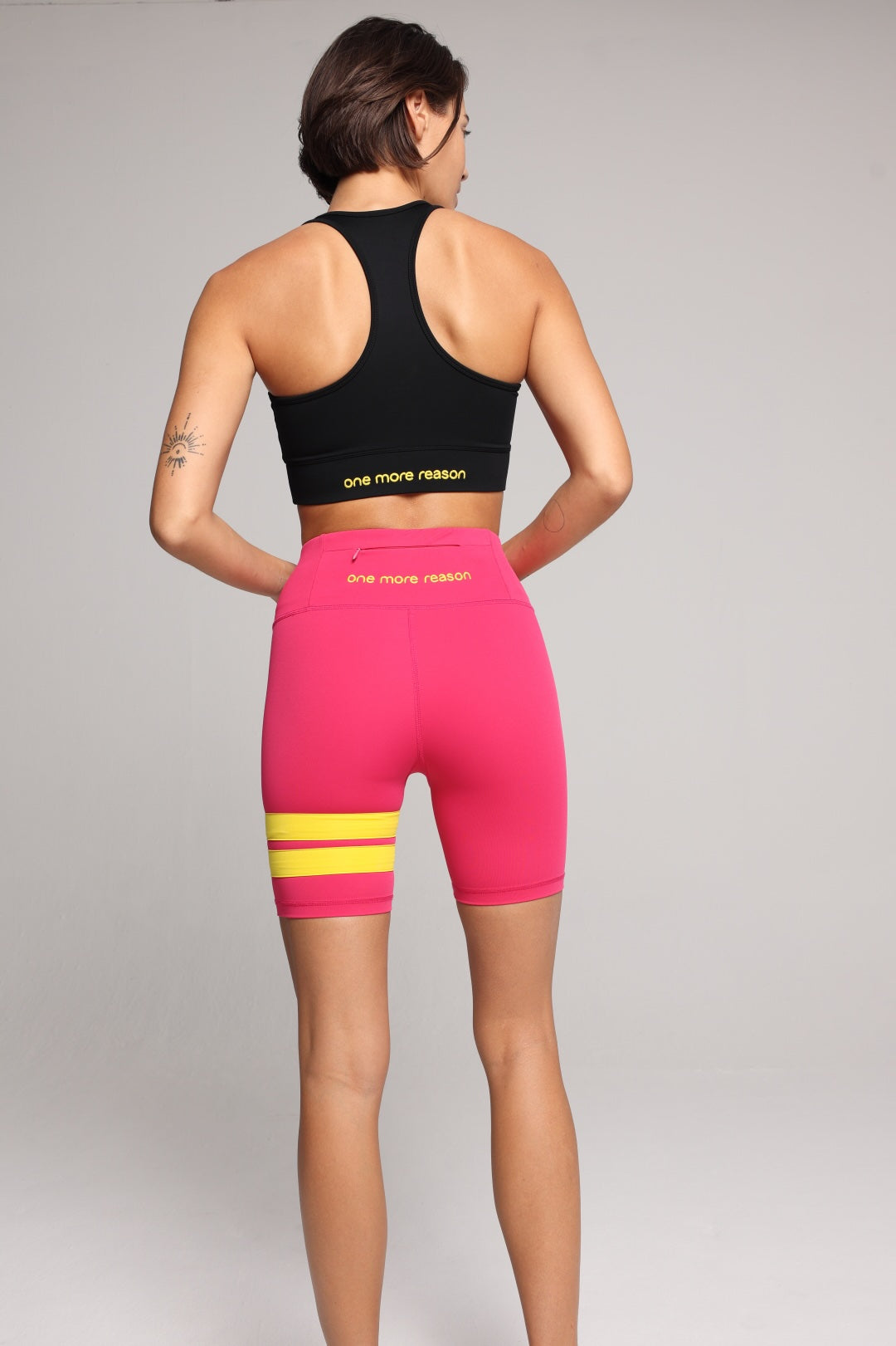A woman shown from the back wearing bright pink athletic biker shorts with yellow horizontal stripes and a black sports bra with yellow text, photographed against a light gray studio background. Premium Sportswear by One More Reason.