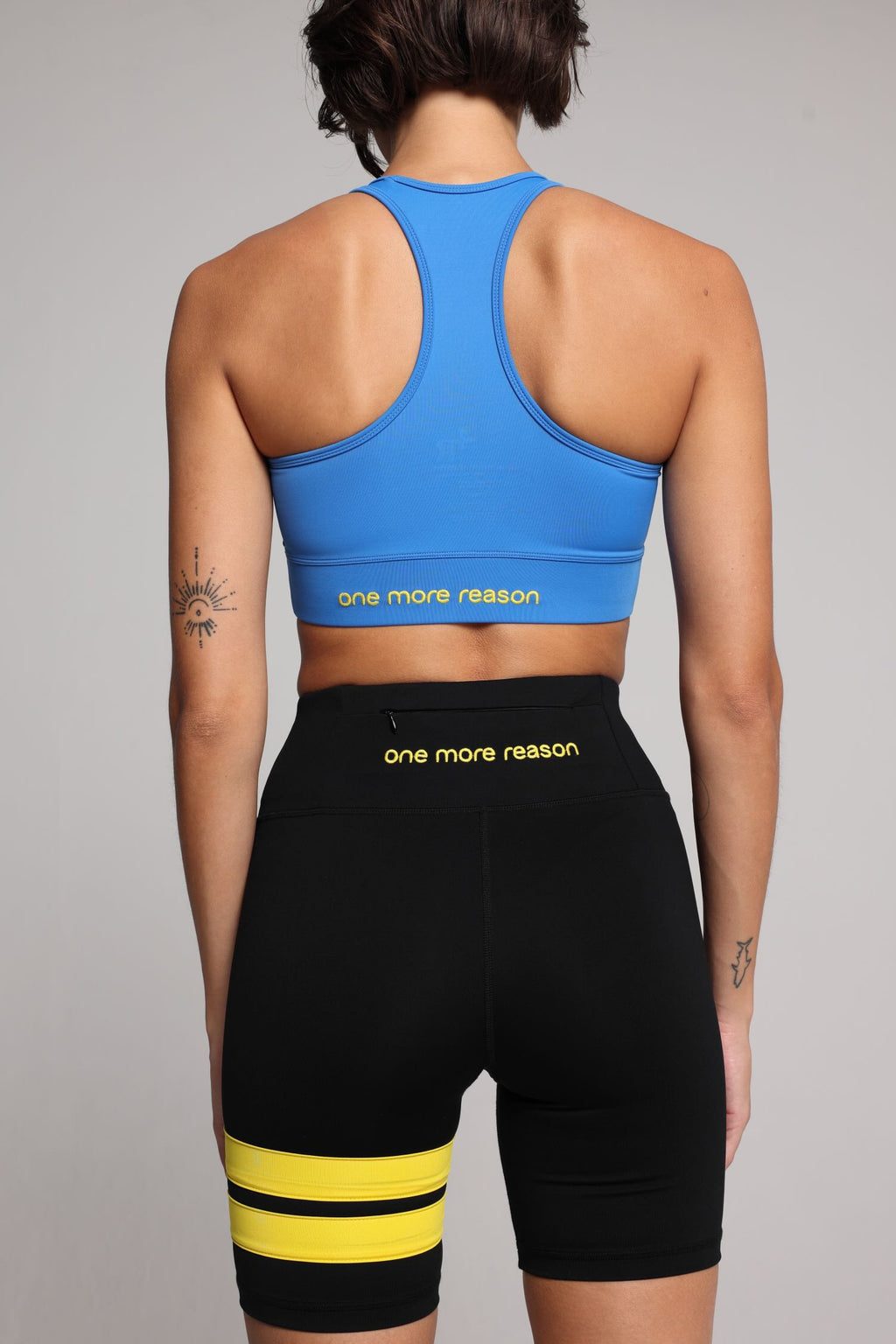 woman in blue sports tank and short black tights with yellow stripes top premium luxury activewear by One More Reason high-performance fitness apparel, female athlete indoor set back view