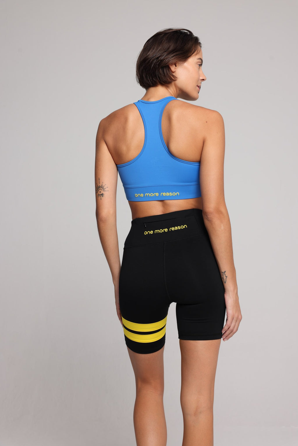 woman in blue sports tank and short black tights with yellow stripes top premium luxury activewear by One More Reason high-performance fitness apparel, female athlete indoor set back view