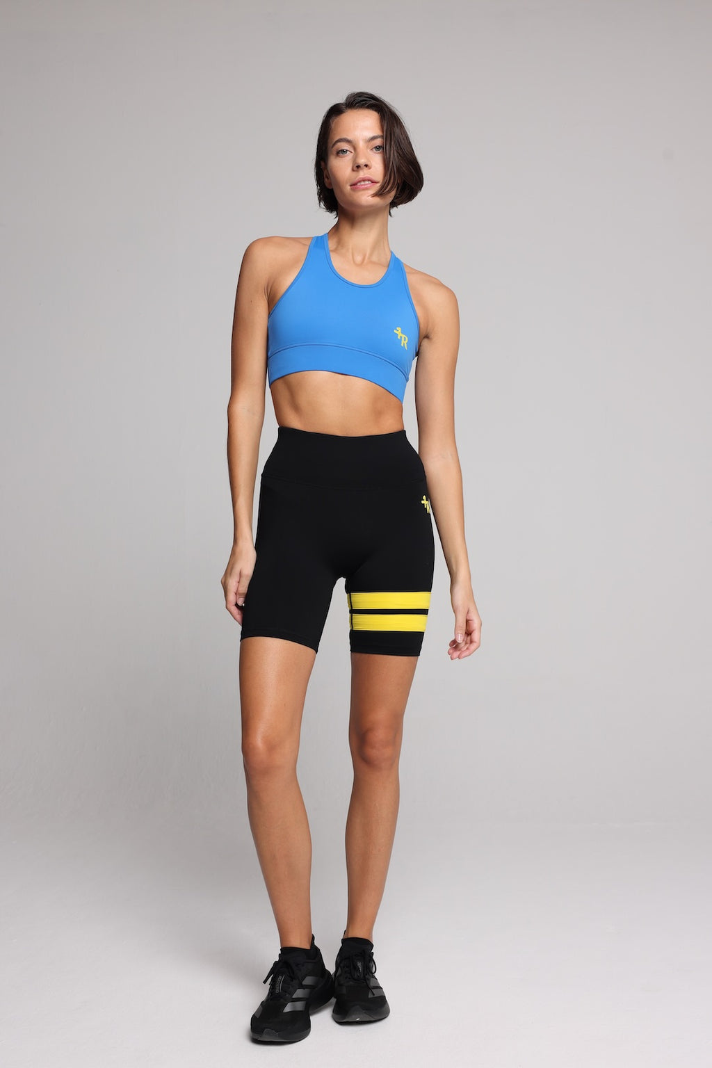 A woman in a bright blue sports bra and black athletic shorts with yellow stripes, standing against a light gray studio background. Premium Sportswear by One More Reason.