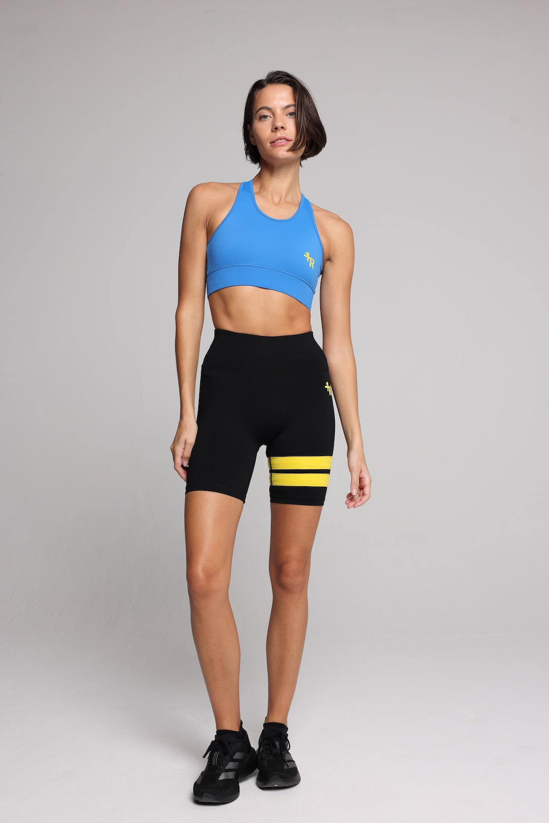 A woman in a bright blue sports bra and black athletic shorts with yellow stripes, standing against a light gray studio background. Premium Sportswear by One More Reason.