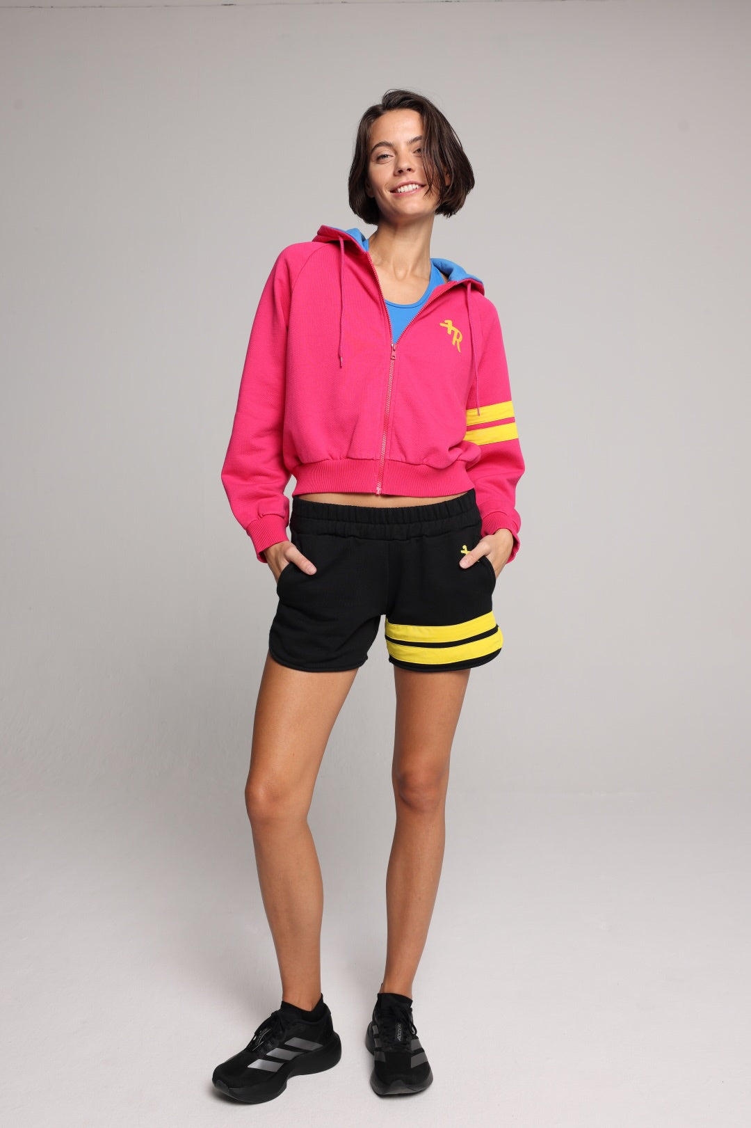 A womanin a bright pink cropped zip-up hoodie with a blue hood lining and yellow sleeve stripes, paired with black athletic shorts featuring yellow stripes and black sneakers, against a light gray studio background. Premium Sportswear by One More Reason.