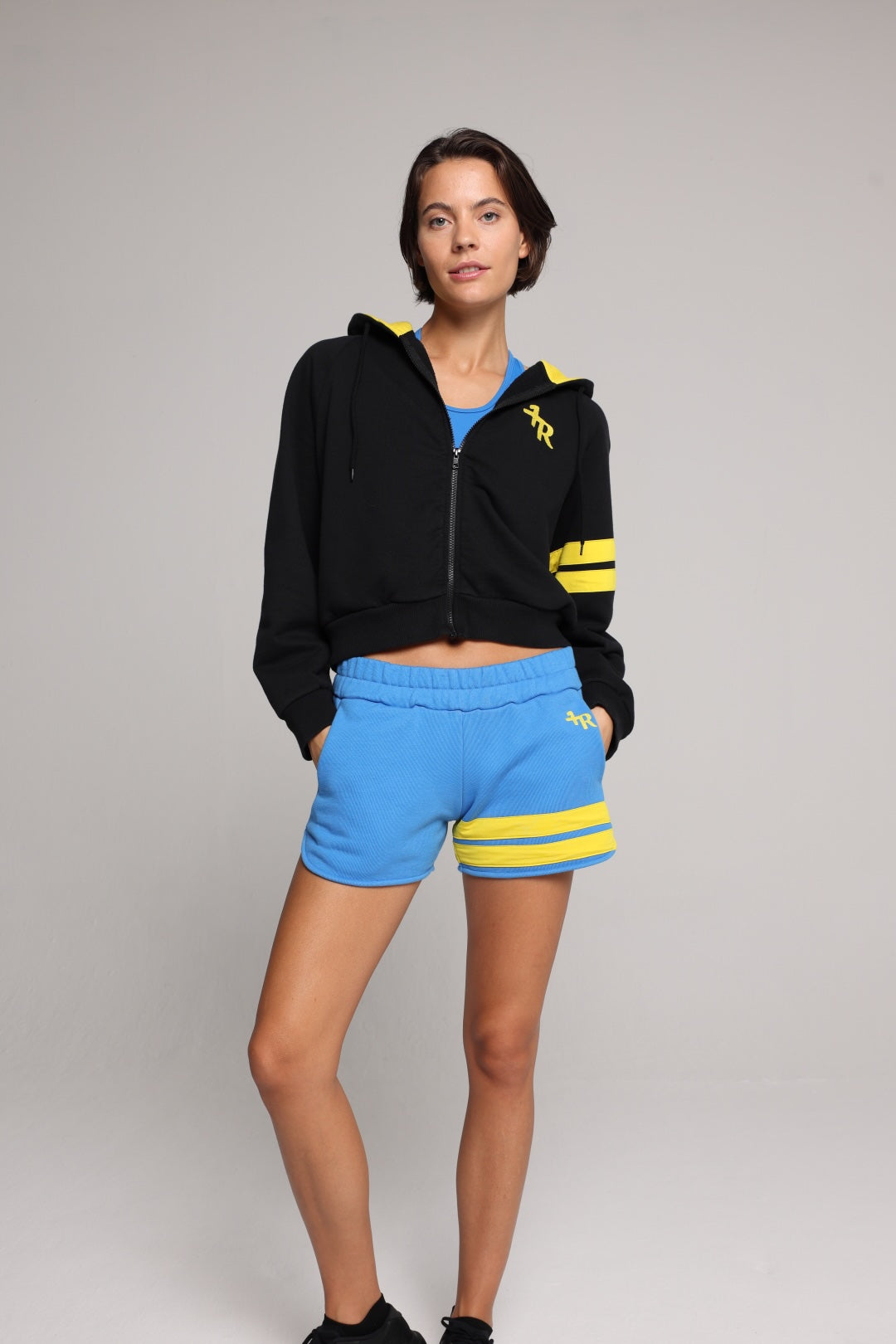 A woman wearing a black cropped zip-up hoodie with yellow stripes, a blue crop top, and bright blue sweat shorts with yellow stripes, standing against a neutral gray studio background. Premium Sportswear by One More Reason.